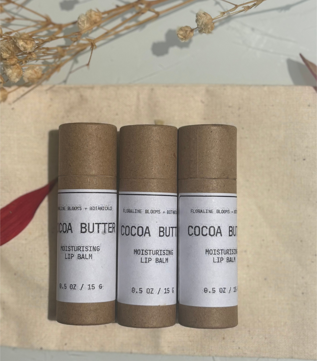 Three cocoa butter lip balm tubes on a wooden surface with dried flowers in the background.